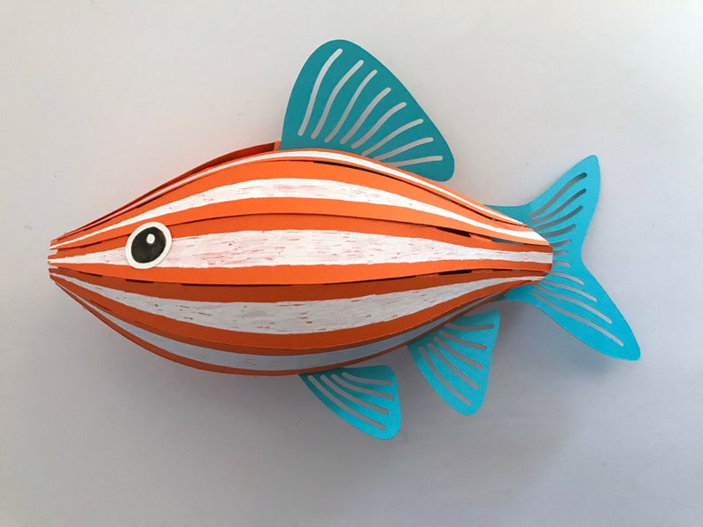 May include: A three-dimensional paper craft fish sculpture featuring alternating horizontal stripes of orange and white. The fish has a rounded body shape with light blue die-cut fins and a tail, and a single black and white circular eye detail.