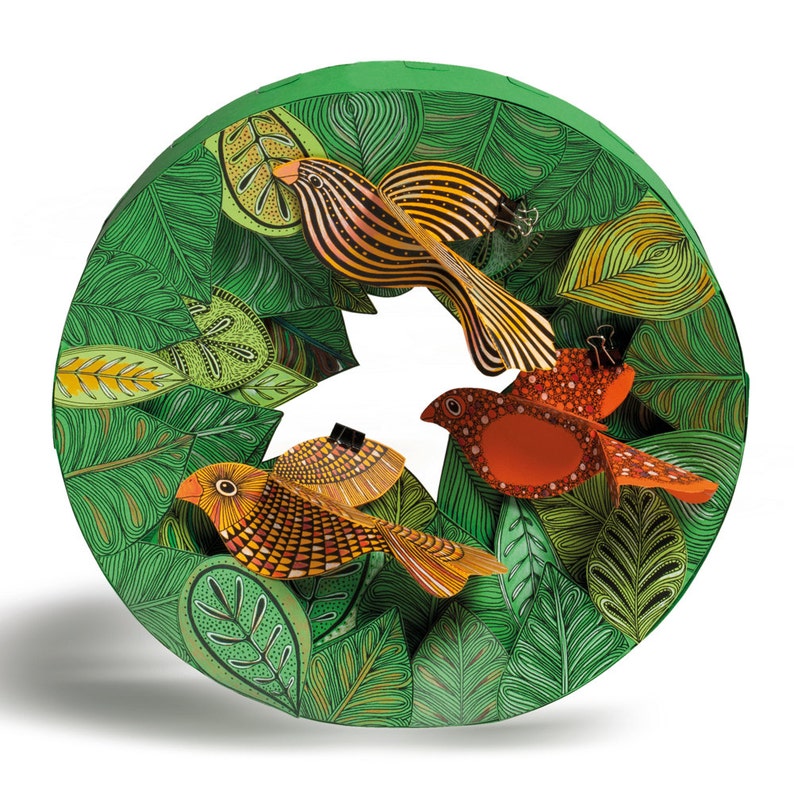 May include: A circular paper craft with a green background and three colorful birds. The birds are orange, red, and yellow with black stripes. The birds are surrounded by green leaves with intricate patterns.