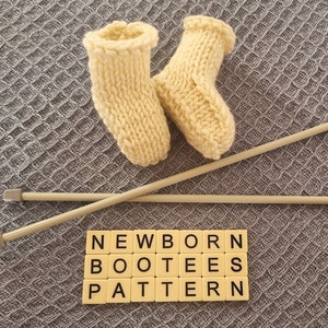 Könnte beinhalten: Ein Paar cremefarbene gestrickte Babyschuhe mit Holzstricknadeln und Buchstabenplättchen, die "NEWBORN BOOTEE PATTERN" buchstabieren.