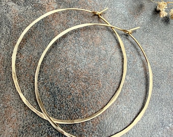May include: A pair of large, circular hoop earrings crafted from thin, hammered gold-tone metal. The earrings feature a delicate, irregular texture along the surface of the wire and include a simple hook-and-loop closure mechanism at the top.