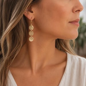 May include: A gold-toned drop earring featuring three vertically aligned circular discs with a textured finish. The earring hangs from a simple hook wire, creating a linear design that rests against the side of the face. The metal has a matte appearance.