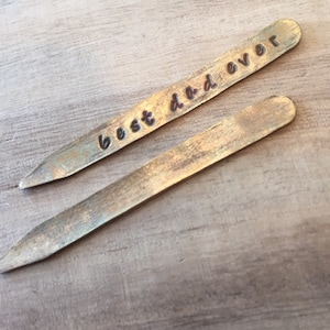 Op de afbeelding: Twee gouden metalen kraagstangen, waarvan één is gegraveerd met de woorden "best dad ever".
