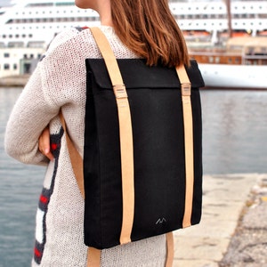 Woman, who is wearing a black backpack with leather straps, is standing on the pier of the port of Trieste. She is facing the sea and a cruise ship. The bag is simple and elegant.