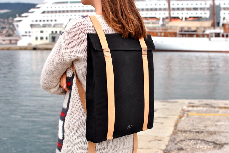 Woman, who is wearing a black backpack with leather straps, is standing on the pier of the port of Trieste. She is facing the sea and a cruise ship. The bag is simple and elegant.