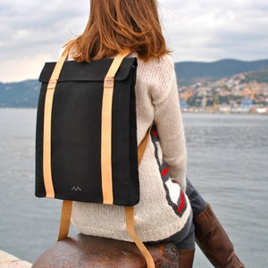 Woman, who is wearing a black backpack with leather straps, is sitting on the pier of the port of Trieste. She is facing the sea. The bag is simple an stylish, the leather straps wrap around the whole bag, making it anti-theft.