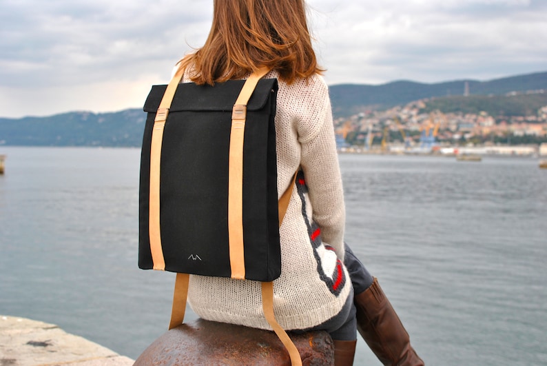 Woman, who is wearing a black backpack with leather straps, is sitting on the pier of the port of Trieste. She is facing the sea. The bag is simple an stylish, the leather straps wrap around the whole bag, making it anti-theft.