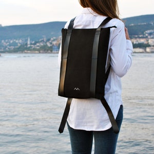 Woman in white shirt is standing in front of the sea and is wearing a full black canvas laptop backpack with black leather straps. The straps wrap around the entire backpack, making it theft-proof.