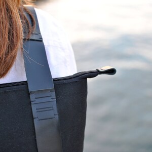 Details of a minimalist hand-stitched black backpack worn by a brown haired woman in white shirt. in the background there is sea water.