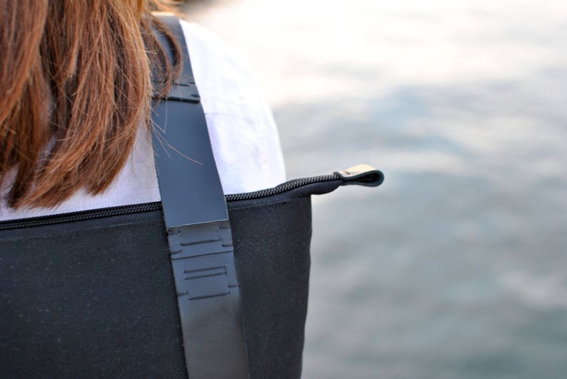 Details of a minimalist hand-stitched black backpack worn by a brown haired woman in white shirt. in the background there is sea water.