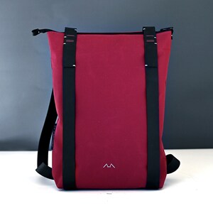 Bordeaux colour minimalist backpack with black leather straps is standing on a white surface in front of a gray background