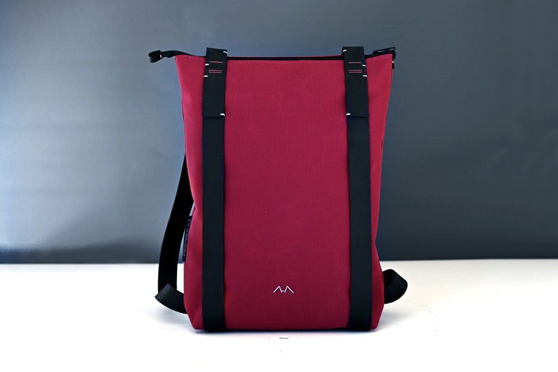 Bordeaux colour minimalist backpack with black leather straps is standing on a white surface in front of a gray background
