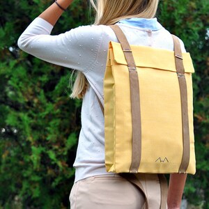Woman, who is wearing a yellow backpack with leather straps, is standing in front of a tree in a garden. The bag's shape is rectangular which is very elegant, but still, the yellow canvas is quite daring and funky.