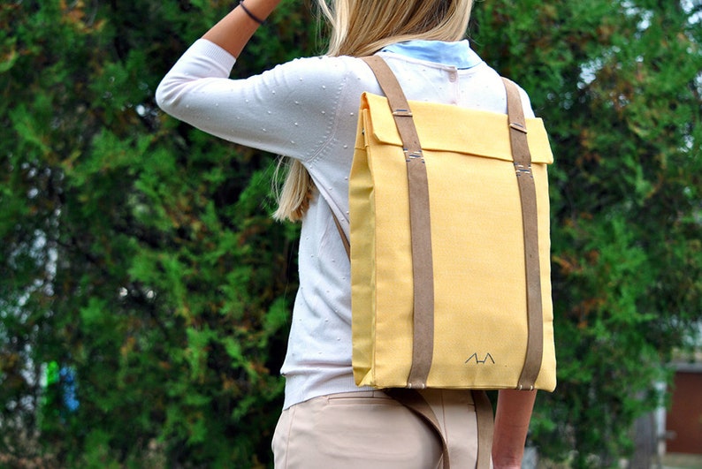 Woman, who is wearing a yellow backpack with leather straps, is standing in front of a tree in a garden. The bag's shape is rectangular which is very elegant, but still, the yellow canvas is quite daring and funky.