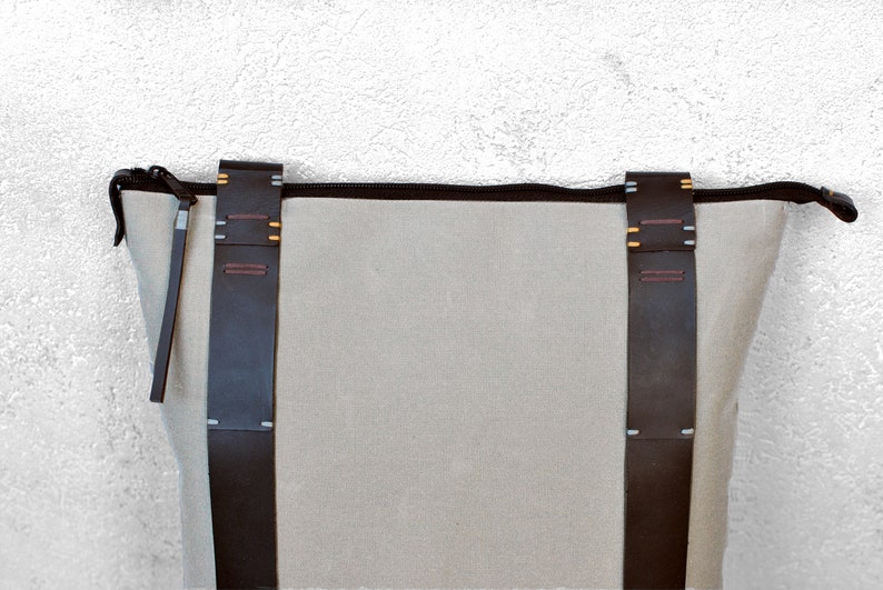 The upper part of a gray canvas minimalist backpack with black zipper and handcrafted leather straps. There are colourful hand-stitched details on the straps. The background is a white wall
