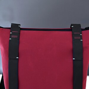 Upper part of a Bordeaux colour minimalist backpack with black zipper and hand stitched black leather straps in front of a gray background