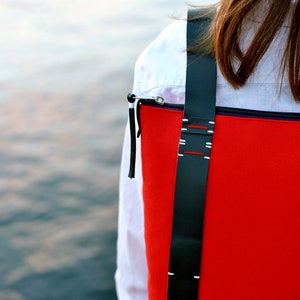 Details of a red minimalist backpack with black leathers hand-stitched straps worn by a woman with long brown hair. She is also wearing a white shirt and standing in front of the water.