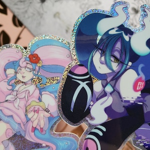 May include: Two holographic stickers featuring anime-style characters. The first sticker features a girl with blue hair and a pink dress. The second sticker features a girl with green hair and a black and purple dress. Both stickers have a glittery finish.