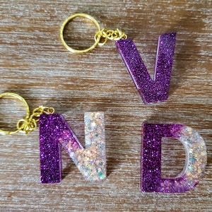 May include: A set of three purple and white glitter resin letter charms on gold keychains. The letters spell "VND".