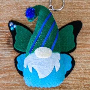 K&ouml;nnte beinhalten: Ein skurriles Wichtel-Schl&uuml;sselanh&auml;nger mit Schmetterlingsfl&uuml;geln. Der Wichtel hat einen gr&uuml;nen Hut mit blauen Streifen und einen blauen Bommel. Die Fl&uuml;gel sind gr&uuml;n mit schwarzen R&auml;ndern und blauen Akzenten. Der Wichtel hat einen wei&szlig;en Bart und ein blaues Gesicht.