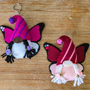K&ouml;nnte beinhalten: Zwei Filz-Wichtel-Schl&uuml;sselanh&auml;nger mit Schmetterlingsfl&uuml;geln. Einer hat einen rosa und lila Hut, der andere einen bordeauxroten und rosa Hut. Beide haben schwarze Z&ouml;pfe, wei&szlig;e B&auml;rte und kleine Rosenakzente. Die Fl&uuml;gel sind schwarz und rosa.