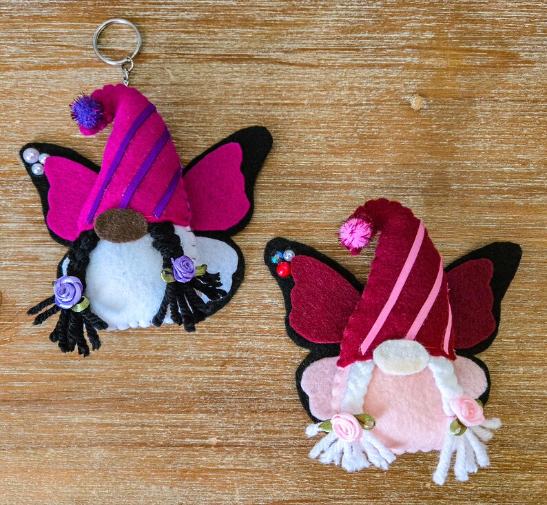 K&ouml;nnte beinhalten: Zwei Filz-Wichtel-Schl&uuml;sselanh&auml;nger mit Schmetterlingsfl&uuml;geln. Einer hat einen rosa und lila Hut, der andere einen bordeauxroten und rosa Hut. Beide haben schwarze Z&ouml;pfe, wei&szlig;e B&auml;rte und kleine Rosenakzente. Die Fl&uuml;gel sind schwarz und rosa.