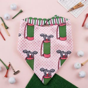 May include: A triangular pet bandana featuring a repeating pattern of green golf bags with pink bases and grey golf clubs on a light pink background with small pink polka dots. The top edge shows a green and white golf tee pattern.