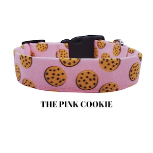 May include: Pink dog collar with a brown and white cookie pattern. The collar has a black buckle and the text "THE PINK COOKIE" is printed below the collar.