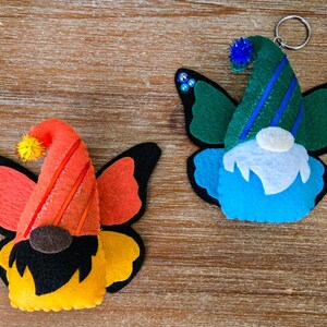K&ouml;nnte beinhalten: Zwei Filz-Schmetterlings-Zwerg-Ornamente. Eines hat einen orangefarbenen Hut mit roten Streifen, orangefarbene Fl&uuml;gel und einen gelb-schwarzen K&ouml;rper. Das andere hat einen gr&uuml;nen Hut mit blauen Streifen, blaue Fl&uuml;gel und einen blauen K&ouml;rper. Beide haben ein Zwerg-Gesicht.