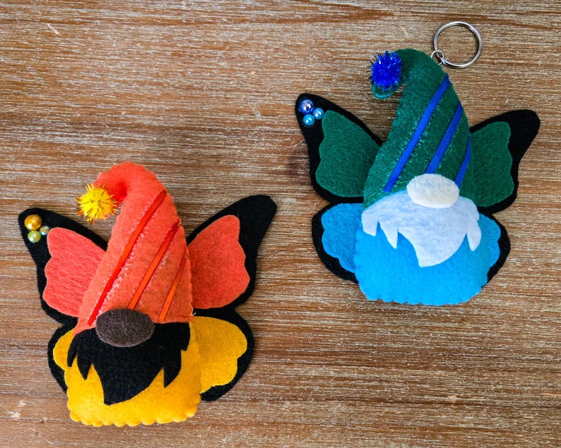 K&ouml;nnte beinhalten: Zwei Filz-Schmetterlings-Zwerg-Ornamente. Eines hat einen orangefarbenen Hut mit roten Streifen, orangefarbene Fl&uuml;gel und einen gelb-schwarzen K&ouml;rper. Das andere hat einen gr&uuml;nen Hut mit blauen Streifen, blaue Fl&uuml;gel und einen blauen K&ouml;rper. Beide haben ein Zwerg-Gesicht.