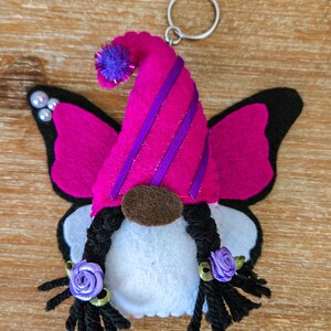 K&ouml;nnte beinhalten: Ein skurriler Wichtel-Schl&uuml;sselanh&auml;nger mit Schmetterlingsfl&uuml;geln. Der Wichtel hat einen rosa Filzhut mit lila Streifen und einem passenden Bommel. Die Fl&uuml;gel sind rosa mit schwarzen R&auml;ndern. Der Wichtel hat schwarze Z&ouml;pfe mit lila Rosenakzenten und eine braune Nase.