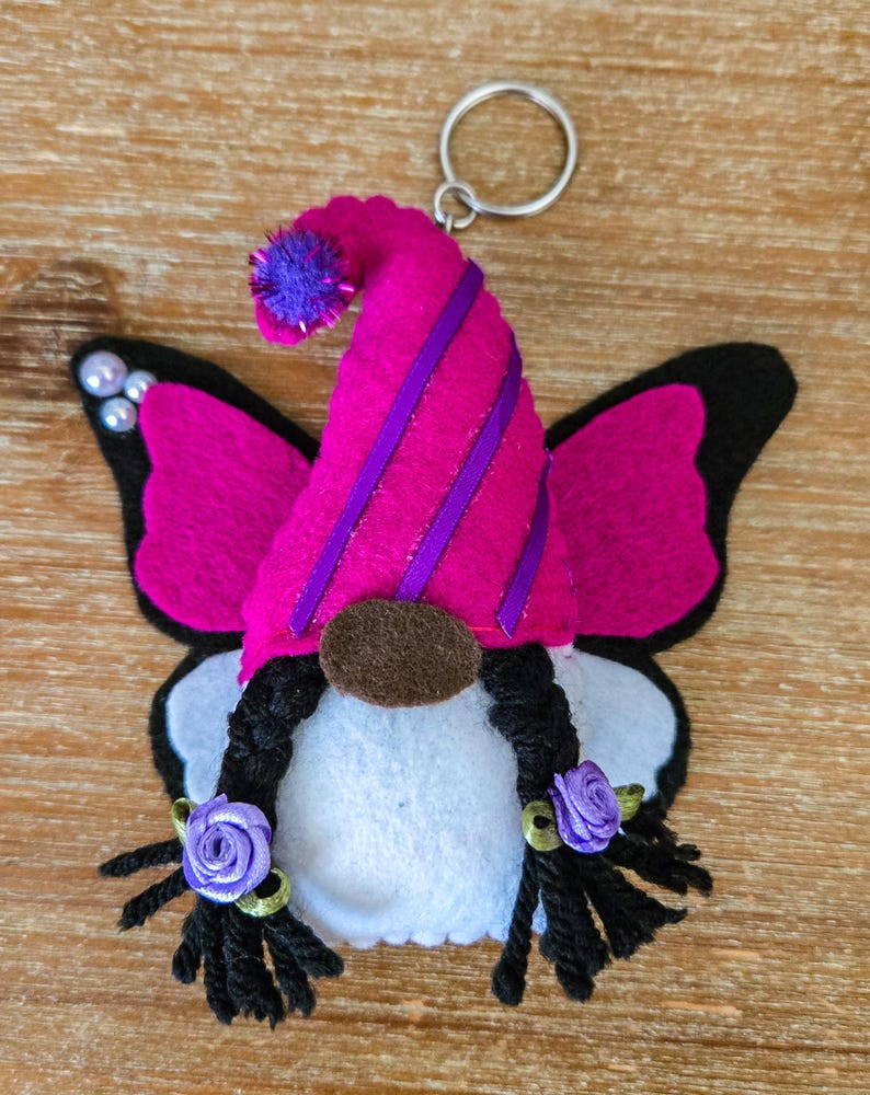 K&ouml;nnte beinhalten: Ein skurriler Wichtel-Schl&uuml;sselanh&auml;nger mit Schmetterlingsfl&uuml;geln. Der Wichtel hat einen rosa Filzhut mit lila Streifen und einem passenden Bommel. Die Fl&uuml;gel sind rosa mit schwarzen R&auml;ndern. Der Wichtel hat schwarze Z&ouml;pfe mit lila Rosenakzenten und eine braune Nase.