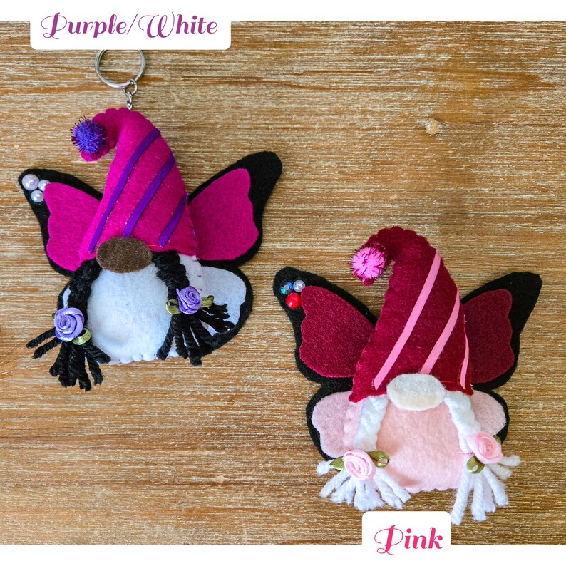 K&ouml;nnte beinhalten: Zwei Filz-Wichtel-Schl&uuml;sselanh&auml;nger mit Schmetterlingsfl&uuml;geln. Einer ist lila und wei&szlig;, der andere rosa. Jeder Wichtel hat eine spitze M&uuml;tze mit Streifen, einen wei&szlig;en Bart und schwarze Z&ouml;pfe. Die Fl&uuml;gel sind schwarz mit rosa Akzenten. Auf einer Holzoberfl&auml;che.