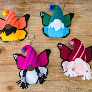 K&ouml;nnte beinhalten: Vier farbenfrohe Filz-Schmetterlings-Gnom-Schl&uuml;sselanh&auml;nger. Jeder Zwerg hat einen andersfarbigen Hut und Schmetterlingsfl&uuml;gel. Die Schl&uuml;sselanh&auml;nger sind orange, gr&uuml;n, rosa und weinrot. Die Gnome haben wei&szlig;e B&auml;rte und einige haben Z&ouml;pfe.