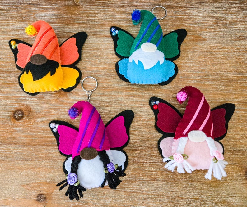 K&ouml;nnte beinhalten: Vier farbenfrohe Filz-Schmetterlings-Gnom-Schl&uuml;sselanh&auml;nger. Jeder Zwerg hat einen andersfarbigen Hut und Schmetterlingsfl&uuml;gel. Die Schl&uuml;sselanh&auml;nger sind orange, gr&uuml;n, rosa und weinrot. Die Gnome haben wei&szlig;e B&auml;rte und einige haben Z&ouml;pfe.
