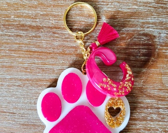May include: Pink and white paw print keychain with a gold heart charm and a pink tassel. The paw print has a white outline and gold glitter accents.