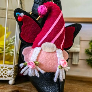 K&ouml;nnte beinhalten: Handgefertigter Zwerg-Ornament mit Schmetterlingsfl&uuml;geln. Der Zwerg hat ein rosa Gesicht, einen wei&szlig;en Bart und einen roten Hut mit rosa Streifen. Die Fl&uuml;gel sind schwarz mit burgunderfarbenen Akzenten. Das Ornament ist an einer schwarzen Ledertasche befestigt.