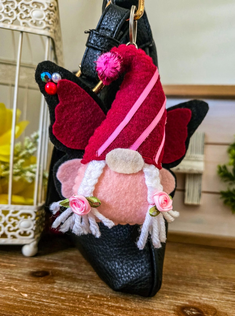 K&ouml;nnte beinhalten: Handgefertigter Zwerg-Ornament mit Schmetterlingsfl&uuml;geln. Der Zwerg hat ein rosa Gesicht, einen wei&szlig;en Bart und einen roten Hut mit rosa Streifen. Die Fl&uuml;gel sind schwarz mit burgunderfarbenen Akzenten. Das Ornament ist an einer schwarzen Ledertasche befestigt.