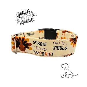 May include: A cream-coloured dog collar with a black buckle and metal hardware. The collar features a repeating pattern of turkeys and the text "Gobble 'Til You Wobble". A festive accessory for pets.