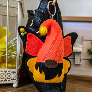 K&ouml;nnte beinhalten: Ein Filz-Wichtel-Schl&uuml;sselanh&auml;nger mit orangefarbenen und schwarzen Schmetterlingsfl&uuml;geln. Der Wichtel hat einen orangefarbenen Hut mit gelben Akzenten und eine braune Nase. Der Schl&uuml;sselanh&auml;nger ist an einer schwarzen Ledertasche befestigt.