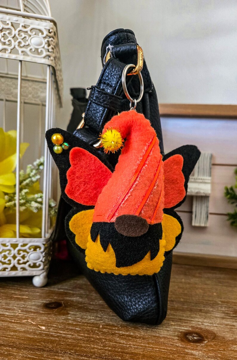 K&ouml;nnte beinhalten: Ein Filz-Wichtel-Schl&uuml;sselanh&auml;nger mit orangefarbenen und schwarzen Schmetterlingsfl&uuml;geln. Der Wichtel hat einen orangefarbenen Hut mit gelben Akzenten und eine braune Nase. Der Schl&uuml;sselanh&auml;nger ist an einer schwarzen Ledertasche befestigt.