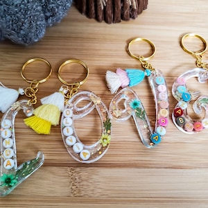 May include: Four clear resin keychains with gold key rings and colorful tassels. Each keychain is shaped like a letter and has a name spelled out with colorful beads. The names are Linda, Olivia, Victor, and Eliza.