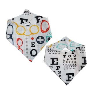 May include: Two white dog bandanas with eye chart and glasses patterns. One bandana features a colorful glasses design with red, yellow, and blue accents, along with the letters "E" and "O". The other bandana displays a classic eye chart.