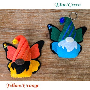 K&ouml;nnte beinhalten: Zwei Filz-Schmetterlings-Zwerg-Ornamente. Eines ist gelb und orange, das andere blau und gr&uuml;n. Jedes hat ein Zwergengesicht, einen Partyhut und Schmetterlingsfl&uuml;gel. Der blau/gr&uuml;ne Zwerg hat einen Schl&uuml;sselanh&auml;nger.