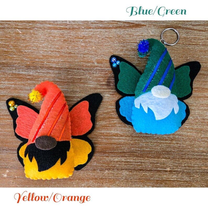 K&ouml;nnte beinhalten: Zwei Filz-Schmetterlings-Zwerg-Ornamente. Eines ist gelb und orange, das andere blau und gr&uuml;n. Jedes hat ein Zwergengesicht, einen Partyhut und Schmetterlingsfl&uuml;gel. Der blau/gr&uuml;ne Zwerg hat einen Schl&uuml;sselanh&auml;nger.