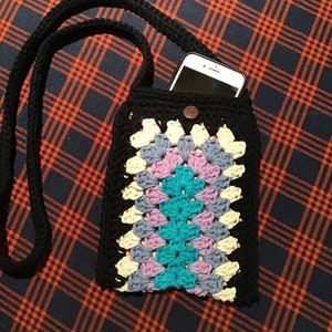 Peut inclure: Une pochette de téléphone en crochet noir avec un motif de granny square coloré. La pochette a une longue sangle noire et une fermeture à bouton. Un smartphone est visible à l'intérieur de la pochette.
