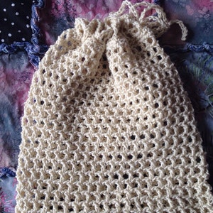 May include: A cream-colored crocheted drawstring bag with a mesh pattern. The bag is lying on a colorful fabric background.