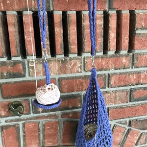 May include: Two handmade blue crochet yarn holders suspended from black hooks against a red brick backdrop. One is a small tray with yarn, the other a larger openwork pouch holding yarn.