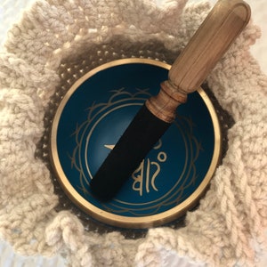 May include: A blue singing bowl with gold trim and a Sanskrit symbol, nestled in a white crocheted bowl. A wooden mallet with a black rubber tip rests on top of the bowl.