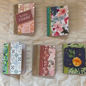 May include: Five small notebooks with different patterned covers. The covers feature floral designs, geometric patterns, and a pineapple design. One notebook has the text "hello there" on the cover.