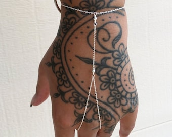 May include: A delicate silver hand chain featuring a thin cable link chain that connects a wrist bracelet to a single finger loop. The jewelry piece is constructed from polished silver metal and consists of a central vertical chain segment.